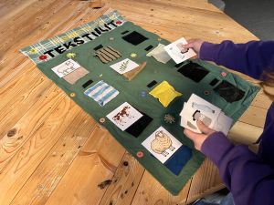 Tekstiilit-peli puisella pöydällä ja kädet asettelemassa kuvia.