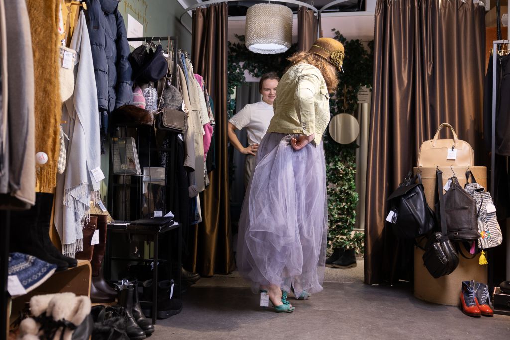 Kaksi henkilöä kokeilee vaatteita. Toisella on hattu ja liila tyllihame, toisella valkoinen t-paita ja harmaat housut. Taustalla sovituskoppeja ja muuta tekstiilirekvisiittaa.